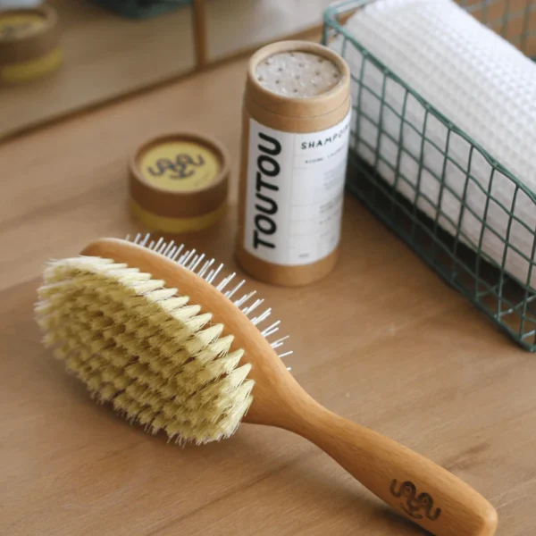 Brosse double face pour chien, en bois naturel, avec un côté en poils doux pour un brossage délicat et un côté en picots métalliques pour démêler efficacement le pelage. Marquée du logo 'TOUTOU' sur le manche.